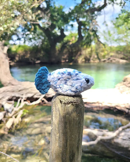 jouet poisson bleu au crochet, texture douce, fait main, bord de l'eau, sur un poteau en bois avec des arbres derrière